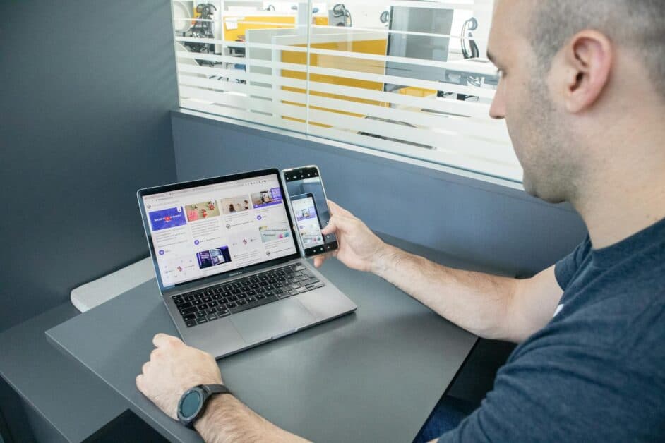Un homme dans un bureau utilisant simultanément un MacBook Pro et un smartphone pour tester l'affichage d'un mur social (Social Wall).