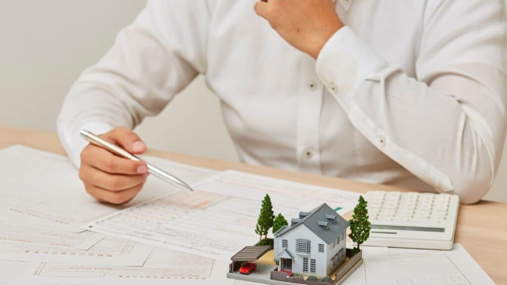 Homme en chemise blanche analysant des documents financiers à côté d'une maquette de maison et d'une calculatrice